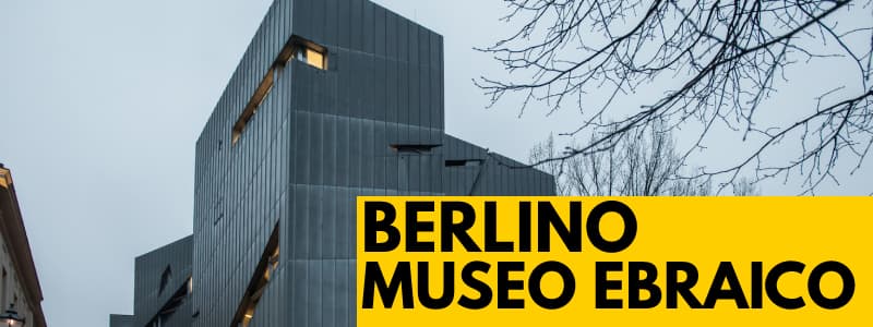 Fotografia dell'esterno del Jüdisches Museum di Berlino in una giornata nuvolosa con rettangolo giallo in basso a destra cons critta nera "Berlino museo ebraico"