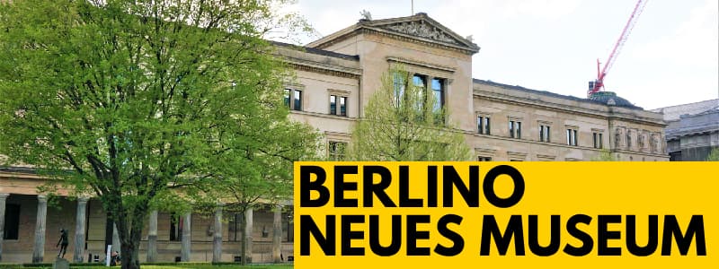 Fotografia della facciata esterna del Neues Museum di Berlino con alcuni alberi in primo piano sulla sinistra. Rettangolo giallo in basso a destra con scritta nera "Berlino Neues Museum"