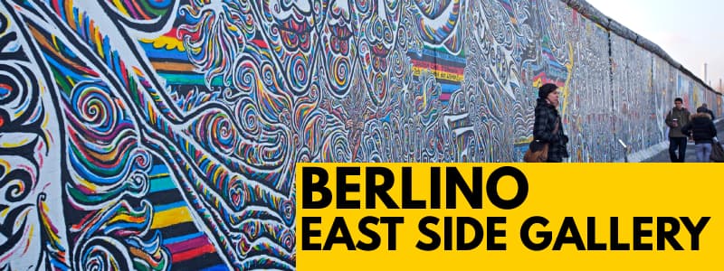 Fotografia di una parte dell'East Side Gallery, l'ultimo residuo del muro di berlino con graffiti e con rettangolo giallo in basso a destra con scritto "Berlino East Side Gallery"